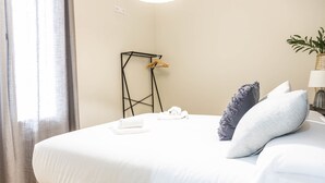 A neatly made bed with white linens, a towel, and a folded towel on the bed. There is a black metal stand with clothes hanging on it. - Tailor s Home by Cadiz Time (Cádiz)