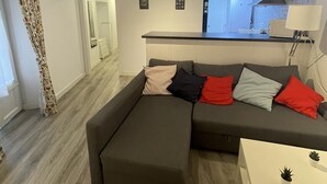 Moderni olohuone, jossa on kulmasohva, pieni valkoinen pöytä ja taustalla keittiötila.