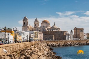 A coastal cityscape with historic architecture, including a prominent building with domes and towers, a rocky waterfront, and a promenade with street lamps. - Casa Rehala Paya by Cadiz Time (Cádiz)
