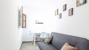 A modern living room with a grey sofa, a white dining table, and a small white chair. There are abstract paintings on the wall and a wooden-framed window. - Casa Rehala Paya by Cadiz Time (Cádiz)