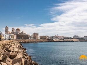 A coastal cityscape with historic buildings, a stone breakwater, and calm sea. - La Roca del Sur by Cadiz Time (Cádiz)
