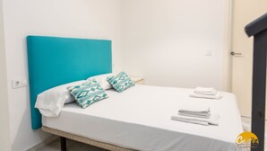 Cama bem arrumada com roupa de cama branca e almofadas verde-água, uma pequena mesa de cabeceira e uma porta à direita.