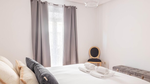 Ein Hotelzimmer mit einem gro ßen Bett, grauen Vorhängen, einem Holzstuhl und einer Bank.