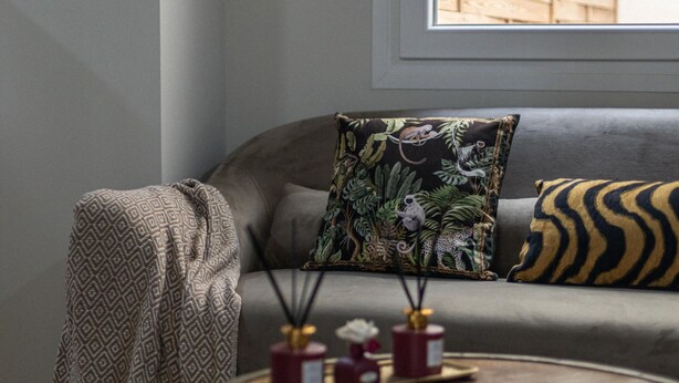 Ett vardagsrum med en grå soffa, en mönstrad pläd, en dekorativ kudde med ett djungeltryck och ett litet bord med två ljus och en vas.