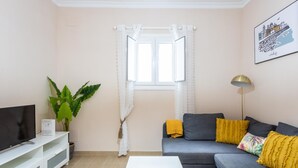 A living room with a grey sofa, a white coffee table, a TV on a white stand, a large plant, and a window with white curtains. - Corralon de Los Carros by Cadiz Time A (Cádiz)