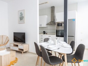 A modern, well-lit living room with a dining area, a kitchen with white cabinets and appliances, and a flat-screen TV. - Apartamento Bike by Livework (Cádiz)