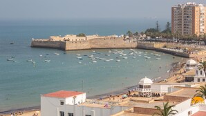A coastal cityscape with a fortified structure, a beach with people, and a modern building with a dome. - Malibu by Cadiz Time (Cádiz)