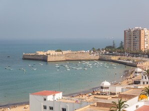 A coastal cityscape with a fortified structure, a beach with people, and a modern building with a dome. - Malibu by Cadiz Time (Cádiz)