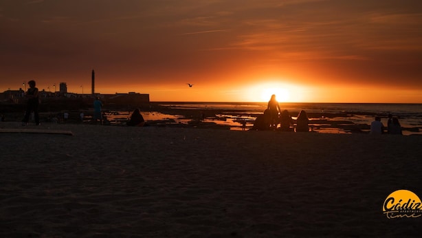 Auringonlasku rannan yllä, jossa näkyy ihmisten hahmoja siluettina ja kauempana oleva rakennus.