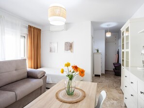 A modern living room with a sofa, a dining table with a vase of flowers, and a kitchen area with cabinets and a hanging light fixture. - San Felipe Neri Free Parking by Cadiz Time (Cádiz)