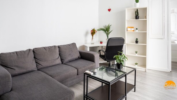 A modern living room with a grey sectional sofa, a glass coffee table, a black office chair, a white desk with a potted plant, and a shelving unit with various items. - Flamenco Centro by Cadiz Time (Cádiz)