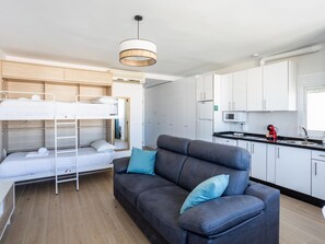 Una habitación moderna con literas, un sofá gris, una cocina con armarios blancos y una lámpara colgante.