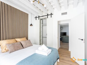 Ein modernes Schlafzimmer mit Bett, einer Wand aus Holzlatten, einer Pendelleuchte und Blick in den Küchenbereich.