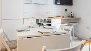 Moderni keittiö, jossa on valkoiset kaapit, seinälle asennettu litteänäyttöinen televisio, joka on sijoitettu työtason yläpuolelle, sekä ruokailutila, jossa on valkoinen pöytä ja neljä astiastoa.