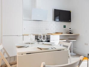 A modern kitchen with white cabinetry, a flat-screen TV mounted above the counter, and a dining area with a white table set for four. - Bajo el Falla by Cadiz Time (Cádiz)