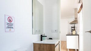 Een moderne badkamer met een houten wastafel, een spiegel en een toilet. Achtergrond is een keuken zichtbaar met een fornuis en kastjes.