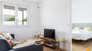 Een moderne woonkamer met een bank, een televisie en een klein tafeltje. In de aangrenzende kamer staat een bed met een groen hoofdeinde.