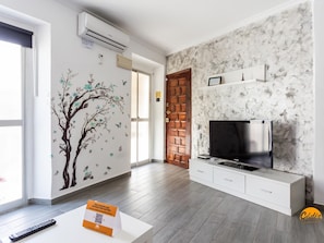 Ein modernes Wohnzimmer mit Fernseher, weißen Schränken, einer Wand mit Marmoroptik und einem Zierbaum-Wandgemälde.