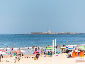A beach with people sunbathing and swimming, colorful umbrellas, and a lighthouse in the background. - Delfin Azul 1 by Cadiz Time (Cádiz)