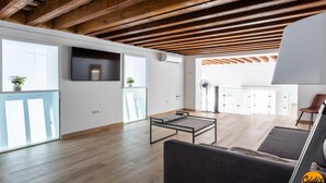 Een ruime woonkamer met houten balken, een bank, een bijzettafeltje en grote ramen.