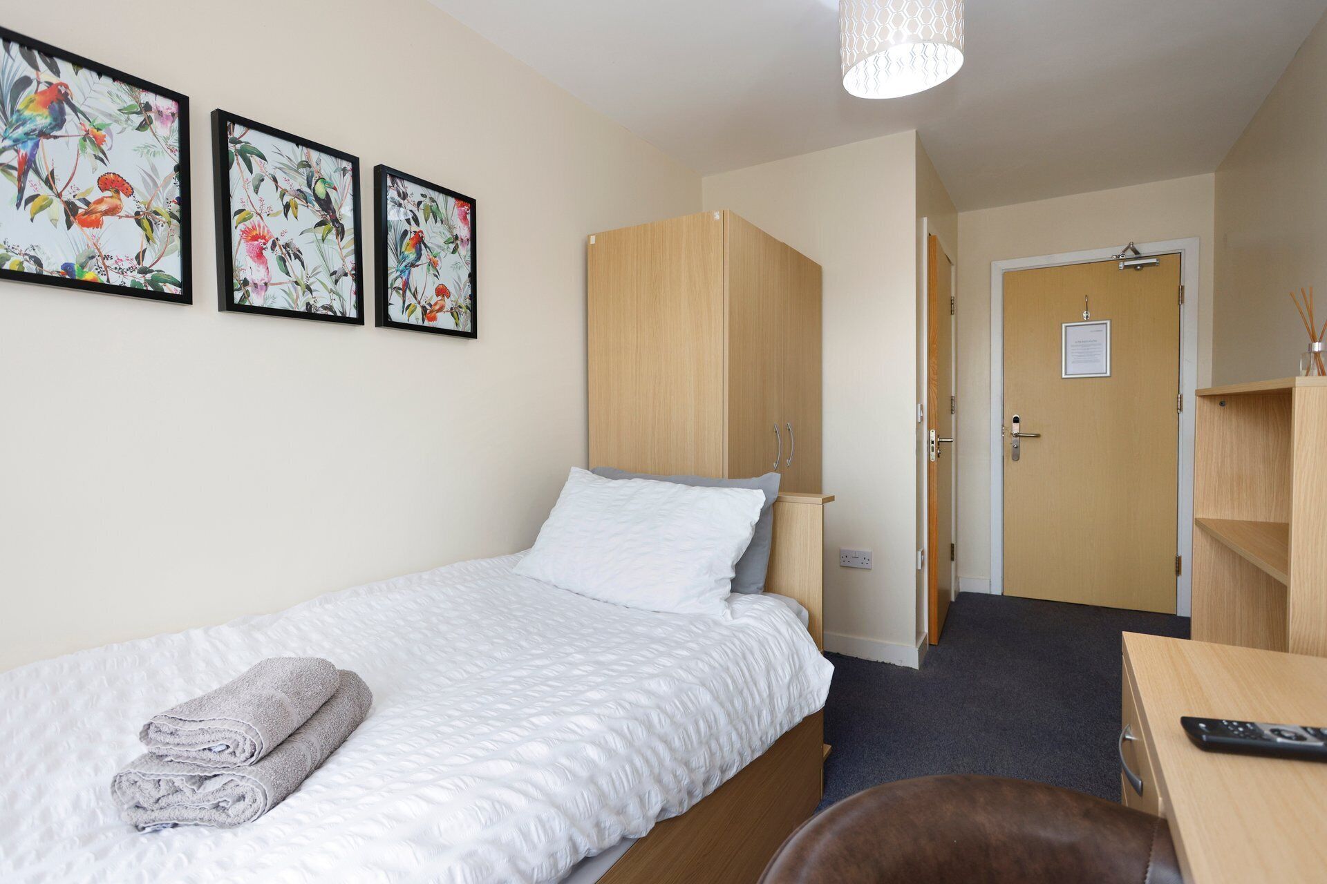 Una cama individual con colcha blanca, un armario de madera, un escritorio con control remoto y dos cuadros florales enmarcados en la pared.