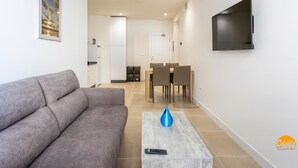 A modern living room with a grey sofa, a coffee table with a remote control, a dining area with a table and chairs, a kitchen with white cabinets, and a TV mounted on the wall. - Delfin Azul 3 by Cadiz Time (Cádiz)