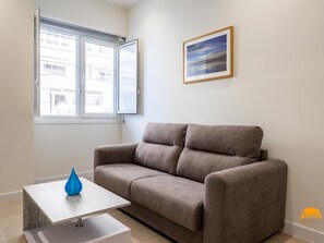 Ein Wohnzimmer mit grauem Sofa, einem weißen Couchtisch und einer blauen Vase. Ein Fenster mit Jalousien befindet sich links, und ein gerahmtes Bild hängt an der Wand.