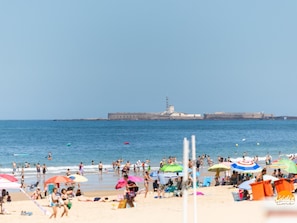 Ein Strand mit badenden Menschen, bunten Sonnenschirmen und einem Leuchtturm im Hintergrund.