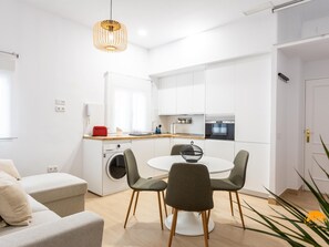 A modern kitchen with white cabinets, a built-in oven, and a red toaster. There is a round dining table with two chairs and a hanging light fixture above. - Brisa de Verano by Cadiz Time (Cádiz)