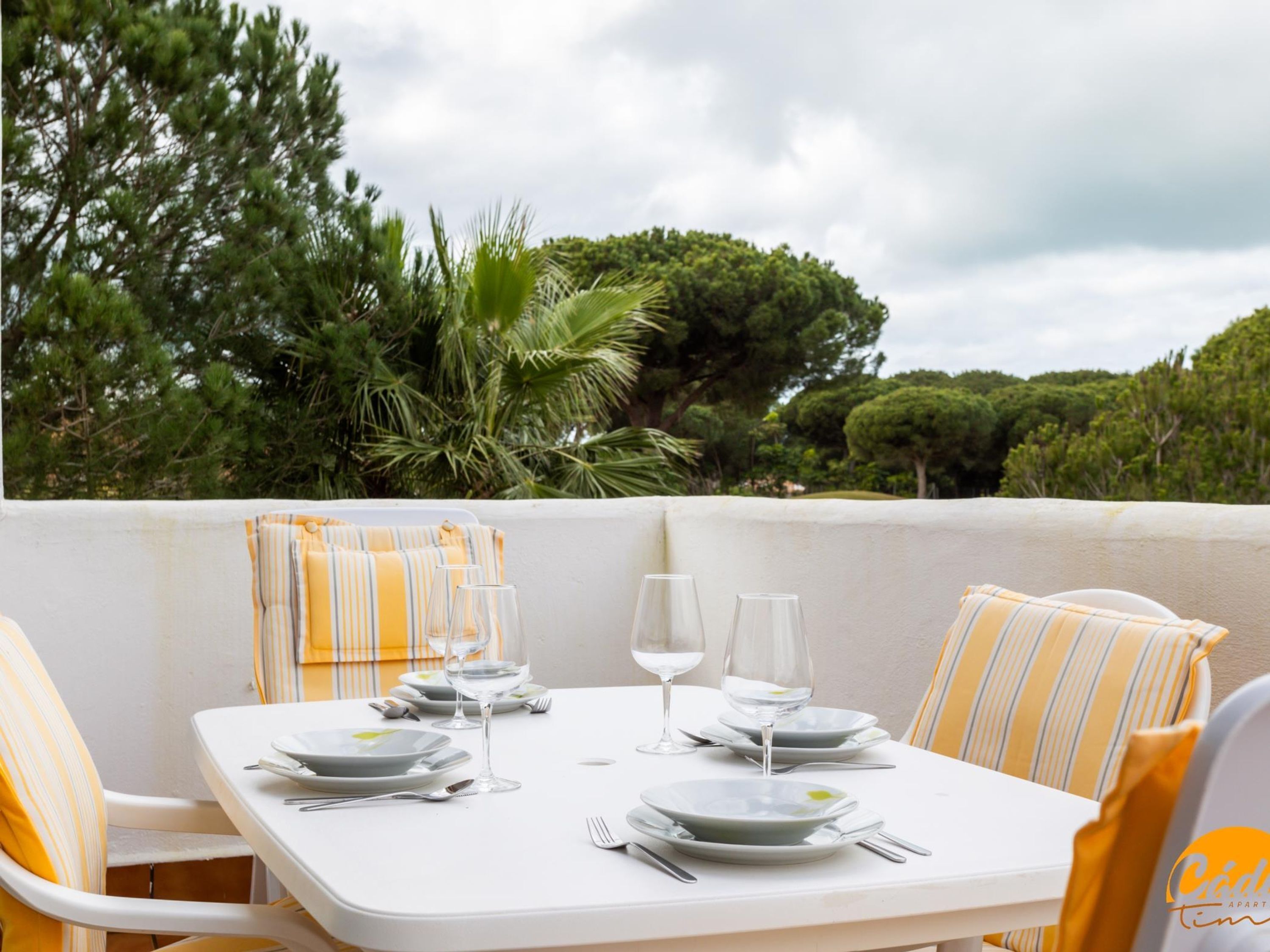 A dining table set for four with white plates, wine glasses, and cutlery, surrounded by striped yellow and white chairs, set against a backdrop of lush greenery and a cloudy sky.