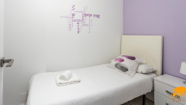 Uma cama bem arrumada com uma toalha branca dobrada, um travesseiro com um desenho roxo e uma pequena mesa de cabeceira com uma luminária.