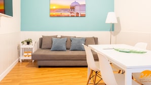 A modern living room with a teal accent wall, a grey sofa with patterned cushions, a white dining table with chairs, and a painting of a sunset over a building. - Apartamento el Patio by C diz Time (Cádiz)