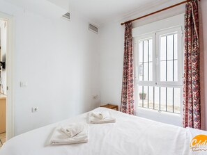A bedroom with a bed, two wall-mounted air vents, a window with a curtain, and a small wooden table. - Apartamento Sol by C diz Time (Conil de la Frontera)