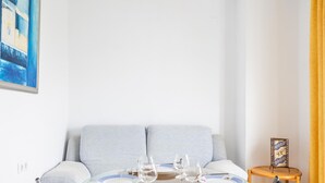 A dining area with a glass table set for four, a sofa, and a small side table with a decorative item. - Apartamento Sue o by C diz Time (Conil de la Frontera)
