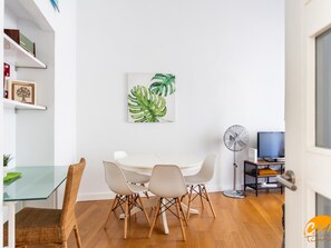 Un coin repas avec une table et des chaises blanches, une œuvre d’art encadrée représentant des feuilles vertes, un ventilateur sur pied et un téléviseur posé sur un meuble.