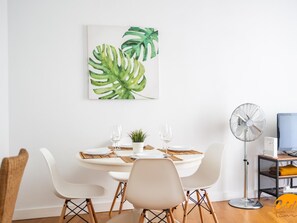 Un coin repas avec une table ronde, des chaises blanches, une petite plante en pot et un ventilateur.