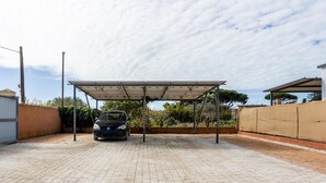 Uma área de estacionamento coberta para carros, com piso pavimentado, um carro estacionado sob uma cobertura e céu claro.
