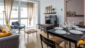 A modern living room with a sofa, TV, dining table set for four, and a large window with curtains. - Teresa Uno by Cadiz Time (Cádiz)