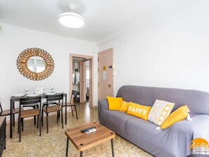 Een woonkamer met een grijze bank, gele kussens, een houten salontafel, en een eetgedeelte met een ronde tafel en stoelen.