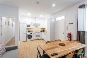 A compact kitchen with a microwave, oven, and washing machine, a wooden dining table with chairs, and a fire extinguisher on the wall. - C diz Living by Livework (Cádiz)