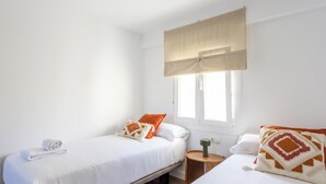 Quarto com duas camas, uma janela com cortina bege, mesa de cabeceira com abajur e toalha na cama à esquerda.