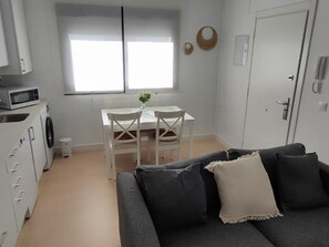 Ett kompakt bostadsutrymme med en grå soffa, ett litet matbord med stolar, en mikrovågsugn och en tvättmaskin.