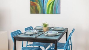 Una mesa de comedor dispuesta con platos, vasos y cubiertos, rodeada de sillas azules, con un centro de mesa con planta y un cuadro abstracto y colorido en la pared.