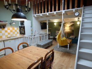 A dining area with a wooden table and chairs, a hanging yellow hammock, a staircase, and a kitchen area with a checkered backsplash. - Entire house for 5 guests –In the heart of the village – Ideal for sports lovers (Saillans)