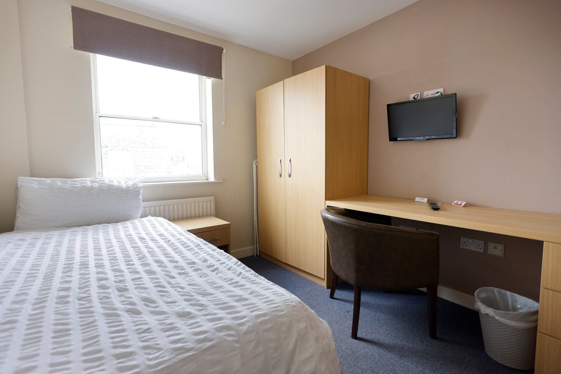 A bedroom with a bed, a desk, a chair, a television, and a window.