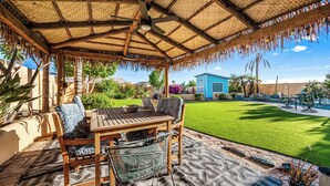 A covered patio with a wooden table and chairs, a thatched roof, and a well-maintained lawn. - SANCTUARY IN THE SUN, 
great location to go or stay (Mesa)