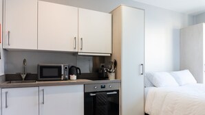 A compact kitchenette with a microwave, kettle, and coffee maker, a small oven, and a bed with white bedding.