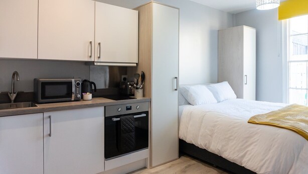 A compact kitchenette with a microwave, toaster, and oven, adjacent to a bed with a white duvet and a yellow throw blanket. - Elegant Studio in Cheltenham (Cheltenham)