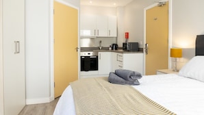 A compact hotel room with a bed, a bedside table, a lamp, a kitchenette with a stove and microwave, and a wardrobe. - Relaxing Retreat in Cheltenham (Cheltenham)