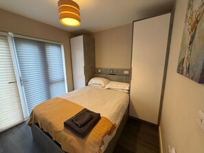 Un dormitorio con cama, mesita de noche, lámpara, ventana con persianas y un cuadro en la pared.